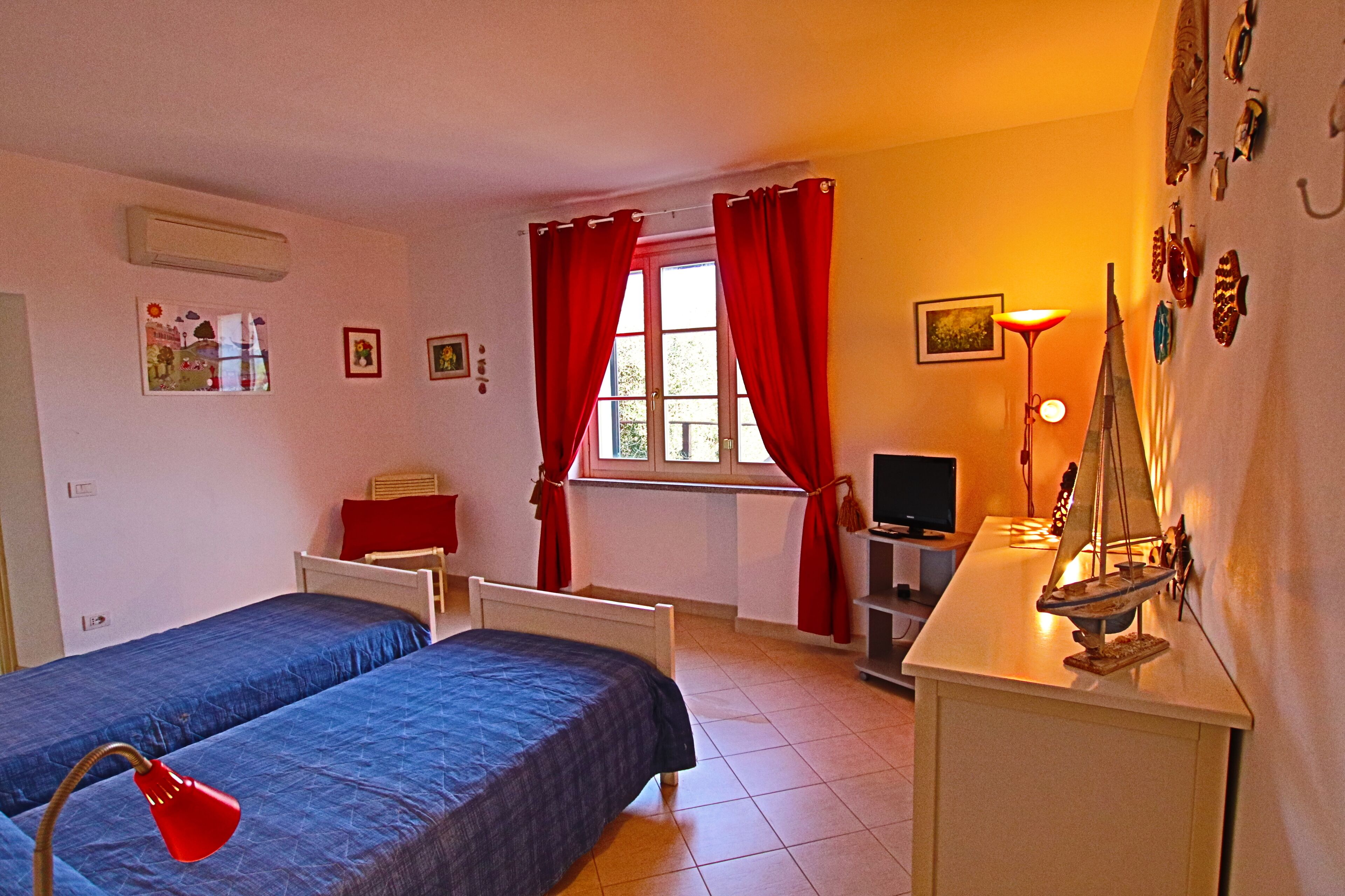 Una camera da letto con due letti, una scrivania con una lampada, una TV e una finestra con tende rosse.
