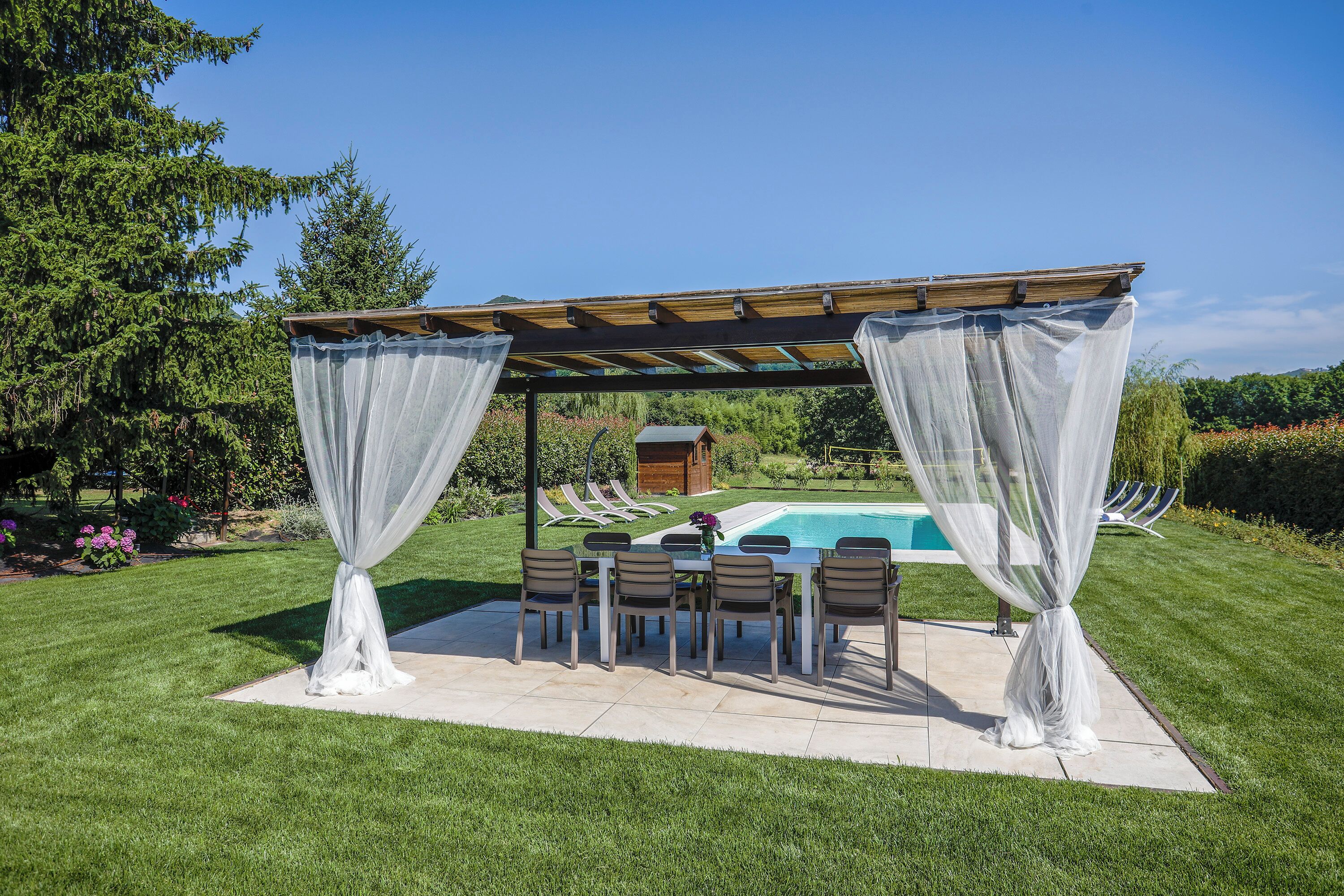 An outdoor patio with a pool, surrounded by a wooden pergola and white curtains.