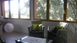Un salón moderno con mobiliario de mimbre, una mesa de centro de cristal y grandes ventanales con vistas a árboles.