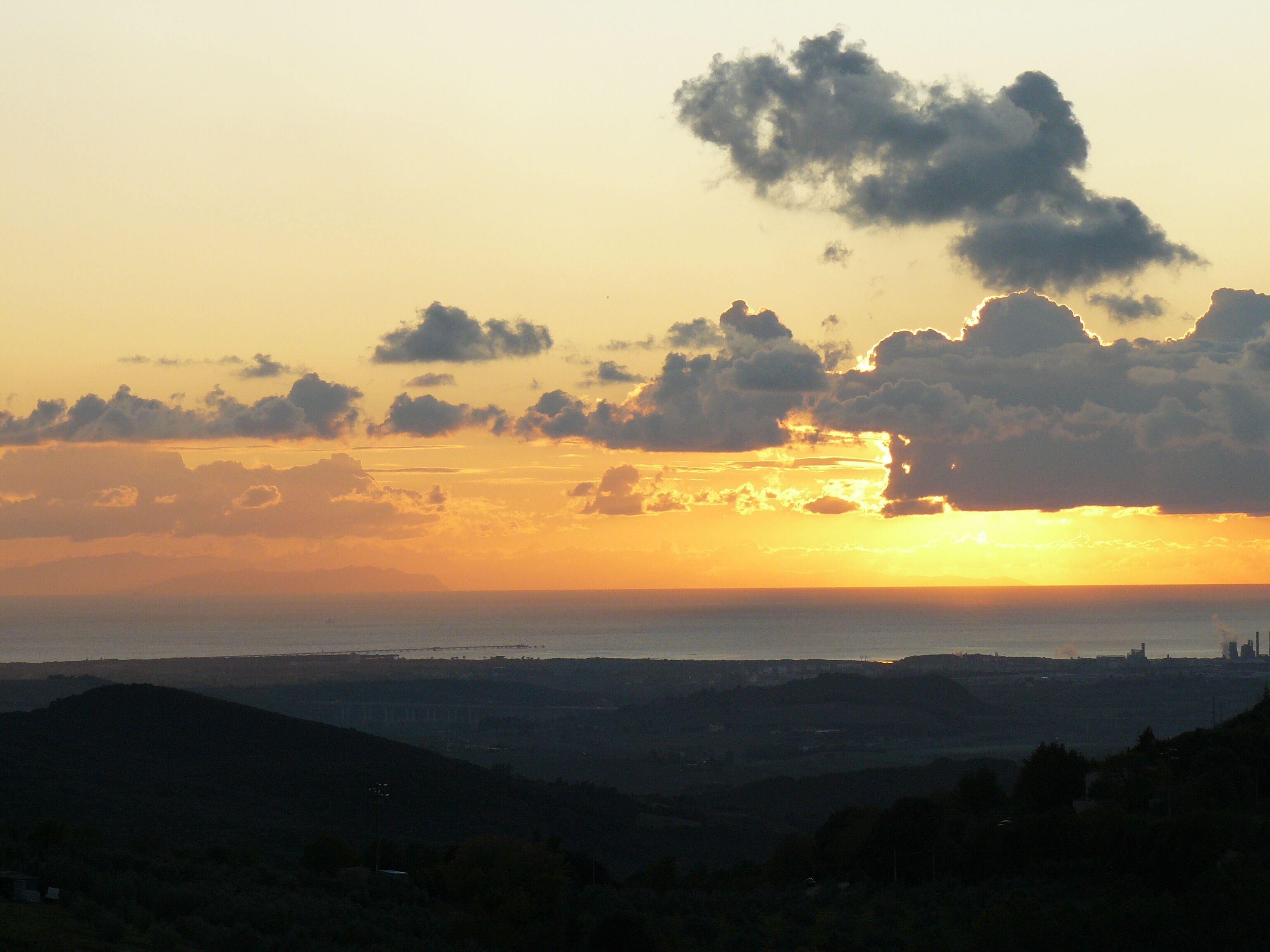 Un coucher de soleil sur un paysage vallonné, avec la silhouette d’une ville lointaine à l’horizon.