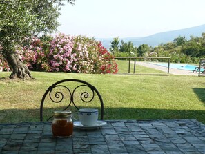 Una mesa con una taza y un tarro, una silla de metal, una piscina y un jardín con flores rosadas y un árbol.
