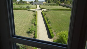 Blick durch ein Fenster auf einen gepflegten Garten mit einem zentralen Weg, ordentlich geschnittenen Hecken und einem Gebäude in der Ferne.