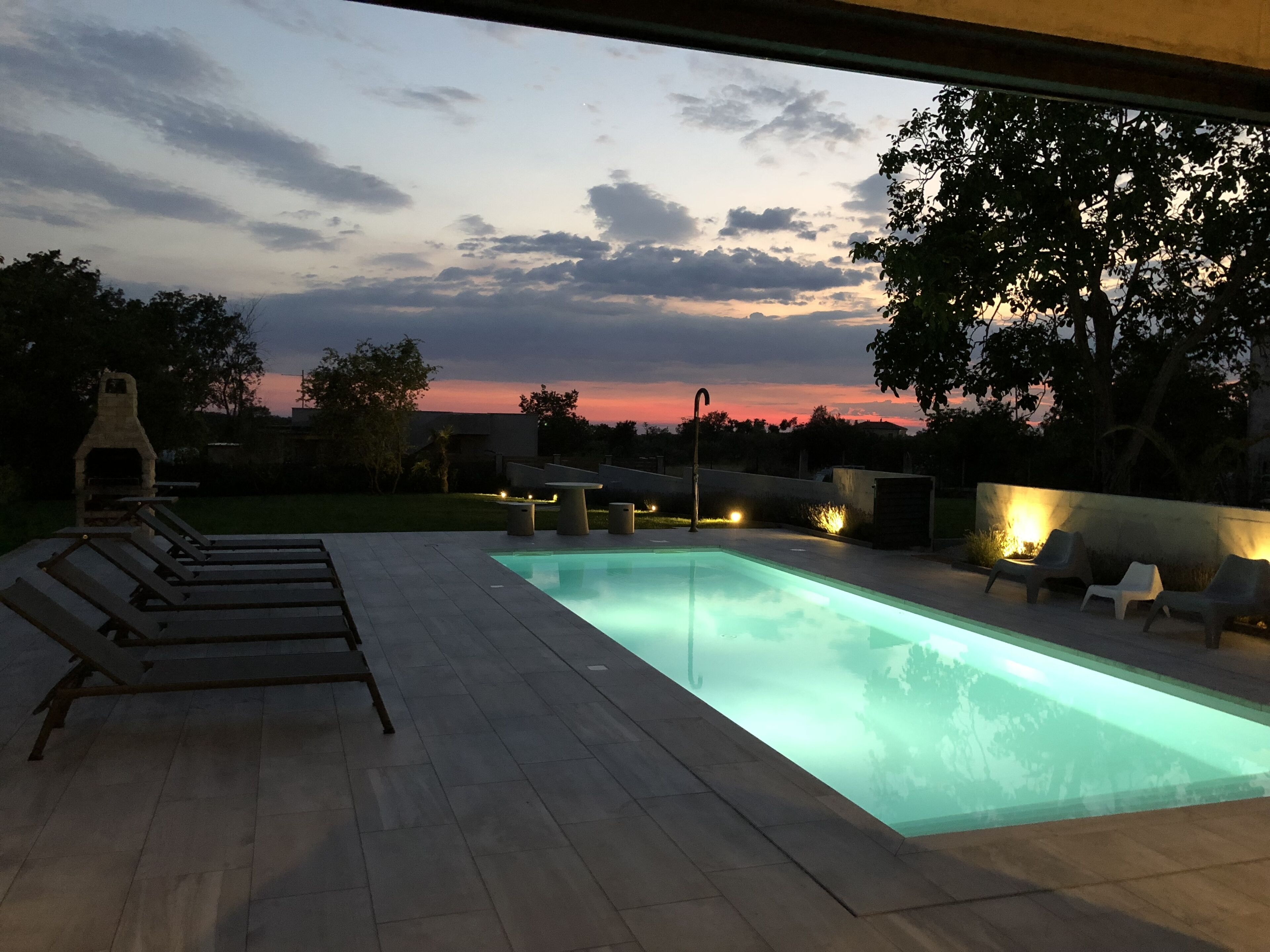 A swimming pool with a view of the sunset, surrounded by lounge chairs and a barbecue grill.