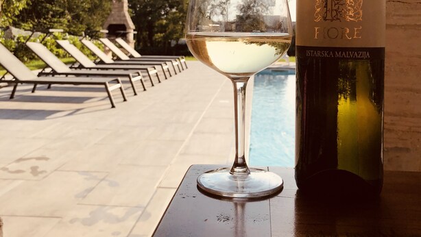 A bottle of Istarska Malvazija wine and a glass of white wine on a table by a poolside with lounge chairs and a stone structure in the background.