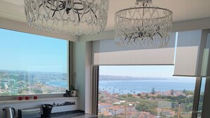 Un comedor moderno con una mesa grande y sillas, una cocina con tostadora y hervidor, y vistas a una ciudad costera a través de amplios ventanales.