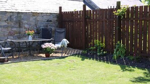Eine Steinsäule, ein Holzzaun, ein Tisch mit Stühlen und Kübelpflanzen in einem Garten.