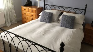 Ein ordentlich bezogenes Bett mit weißer Bettwäsche, zwei grauen Strickkissen und eine Holzkommode mit einer Lampe daneben.