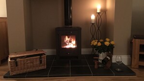 Ein Kaminofen mit sichtbarem Feuer, ein Kerzenhalter mit Kerzen, ein Korb und eine Vase mit Blumen auf einem Fliesenboden.