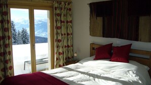 A bedroom with a bed, red pillows, a wooden headboard, a window with curtains, and a view of snow-covered mountains. - Luxury Modern Swiss Chalet - Spectacular Panoramic Views - 200m From Piste (Gryon)