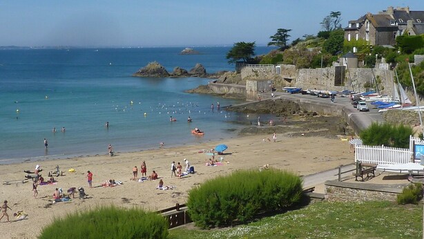 Una playa con gente tomando el sol y nadando, un litoral rocoso y edificios al fondo.
