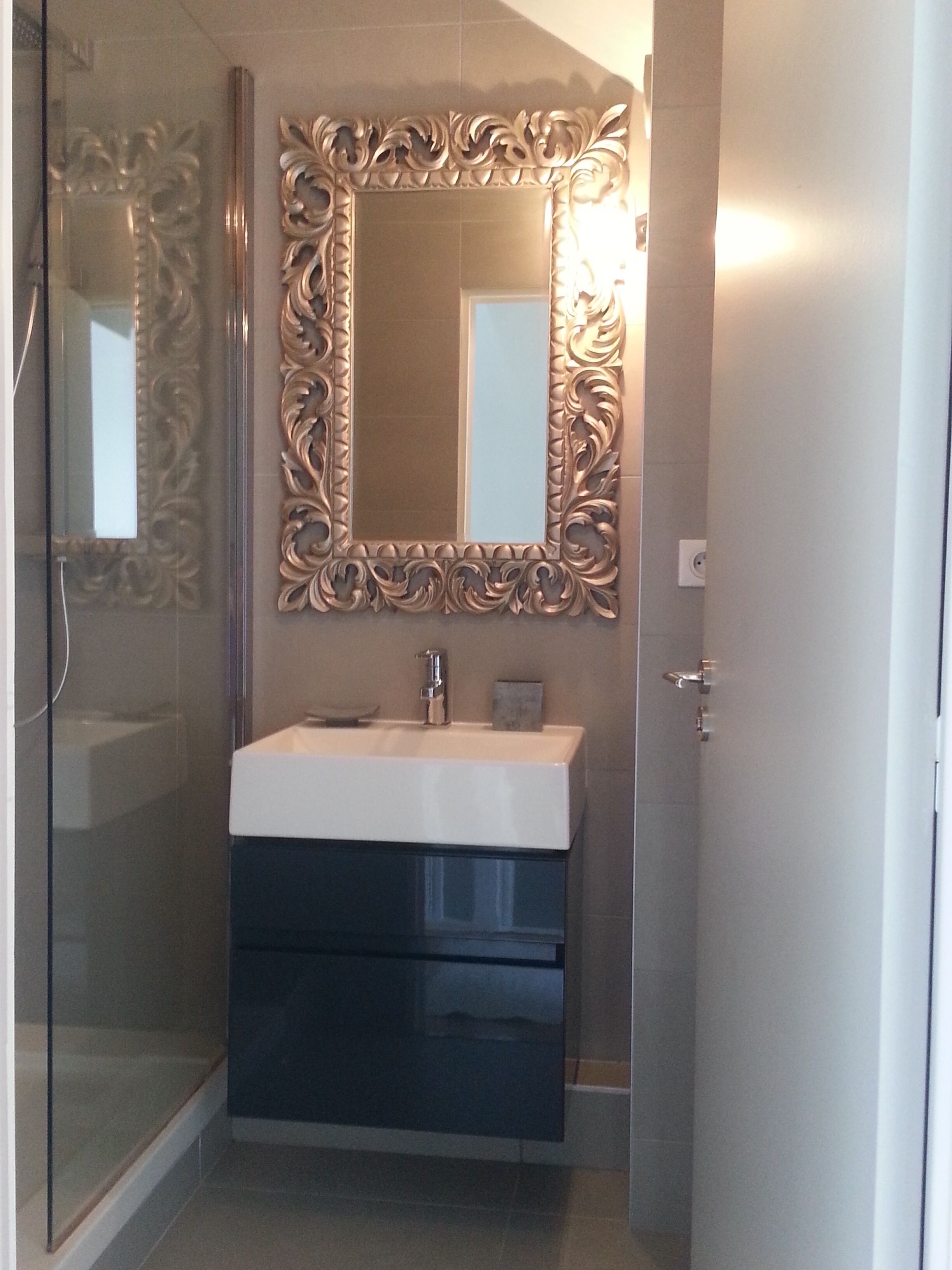 Un baño con cabina de ducha de vidrio, un lavamanos blanco con mueble oscuro y un espejo decorativo con marco ornamentado.