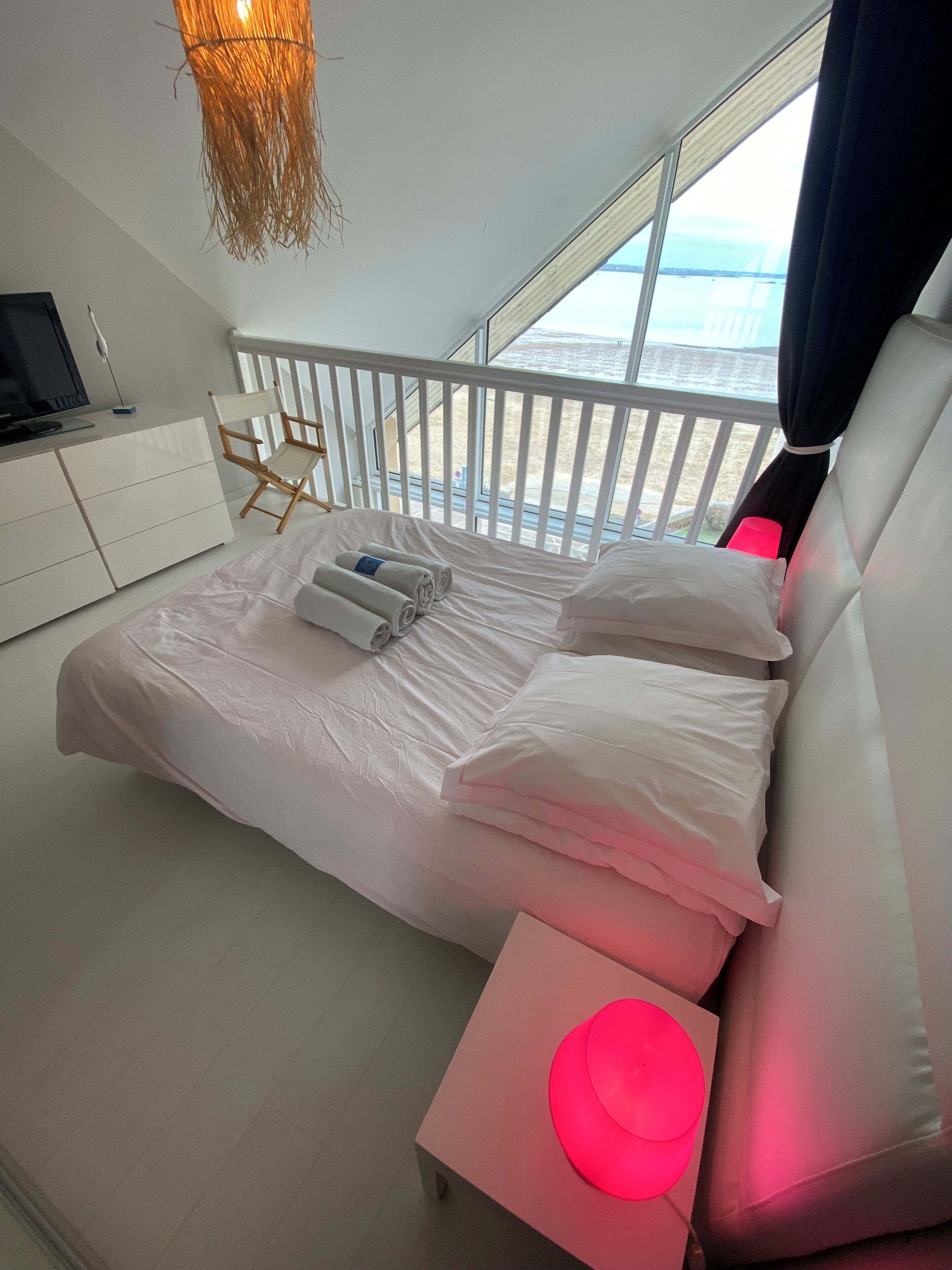 A modern bedroom with a large bed, a bedside table with a red lamp, and a view of the sea through a balcony.