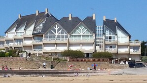 Un edificio frente al mar con varios balcones, una playa arenosa en primer plano y un cielo azul despejado.