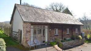 A rustic stone and wooden cottage with a sloped roof, a glass door entrance, and a small garden area with two white chairs. - Lovely Devon barn, near Exeter, set in 15 acres of countryside (Crediton)