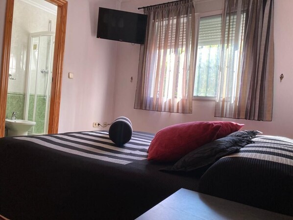 A bedroom with a bed, a TV mounted on the wall, a window with curtains, and a bathroom visible through an open door.