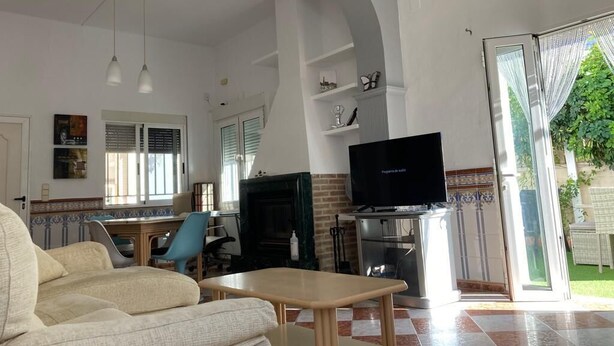 Ein geräumiges Wohnzimmer mit Sofa, Couchtisch, Fernseher und Kamin. Ein Glastür öffnet sich zu einem Garten.