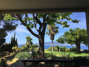 A view from a balcony overlooking a garden with a large tree, a lawn, and a body of water in the distance.