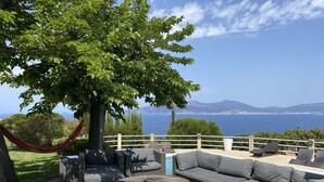 An outdoor seating area with a large tree, a hammock, and a view of the sea and mountains.