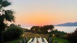 A long dining table set for a meal with chairs on both sides, overlooking a body of water and mountains at sunset.