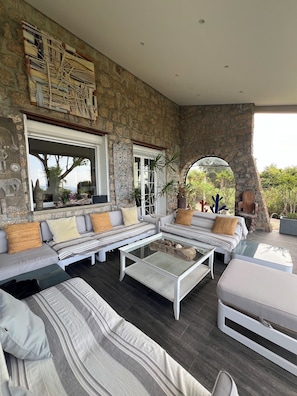 A spacious outdoor seating area with a stone wall backdrop, a large window, and a view of the surrounding landscape. - Superbe villa avec vue exceptionnelle sur le golfe d’Ajaccio.  (Pietrosella)
