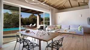 A spacious living area with a wooden table, chairs, and a sofa. A large window offers a view of a swimming pool and trees. - Aggelos Brand New Exclusive Villa with Private Pool (Agrikies Villas) (Lefkada)