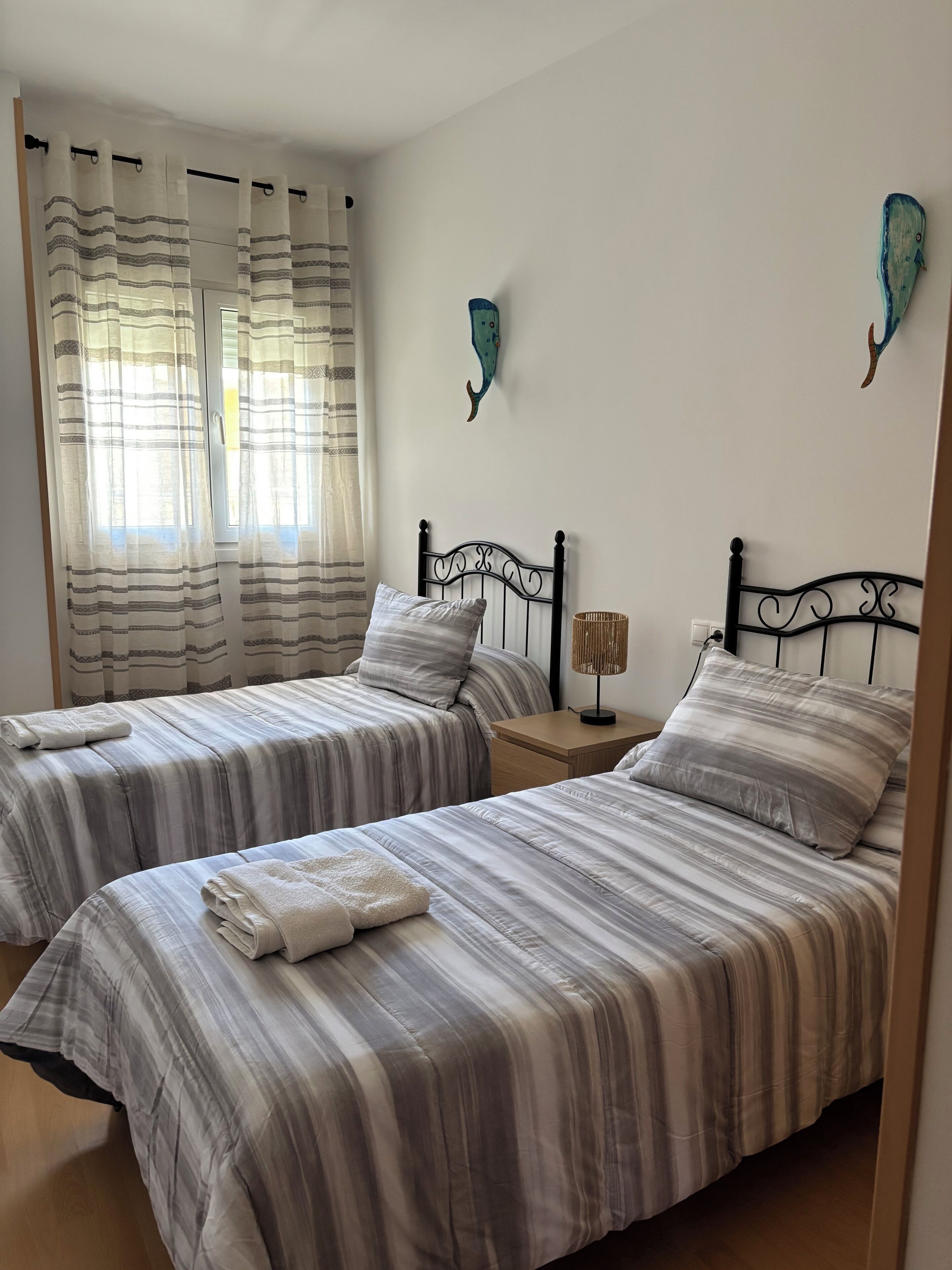 A room with two beds, each with striped bedding and pillows, a bedside table with a lamp, and a wall-mounted decoration.