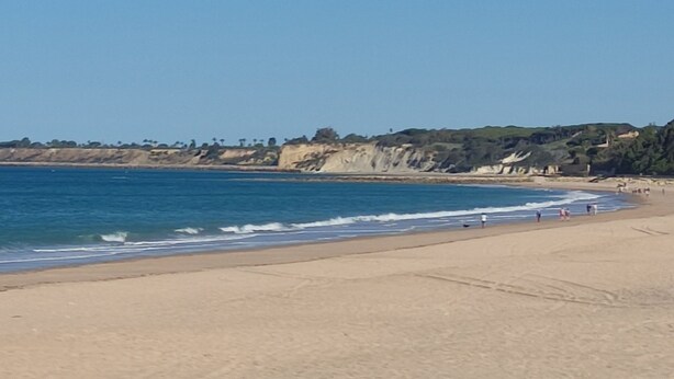Una playa con cielo despejado, orilla arenosa y acantilados rocosos al fondo.