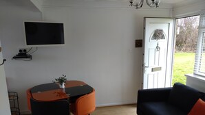 Een ruimtelijk ingerichte leefruimte met een eettafel, een bank, een aan de muur bevestigde televisie en een deur naar buiten.