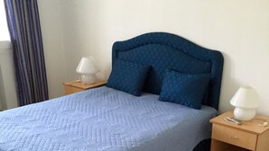 Una cama con colcha azul acolchada y almohadas, flanqueada por dos mesitas de noche con lámparas y un cajón.