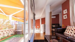 A modern living room with a leather sofa, a coffee table, and a glass door leading to a balcony with a wooden deck and a garden.