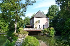 A house with a stone bridge over a river, surrounded by lush greenery.