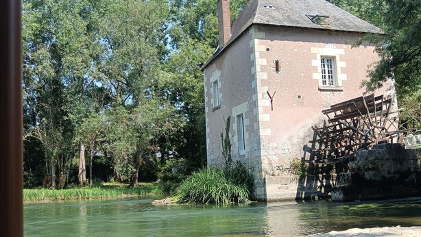 A rustic building by a river with a thatched roof and a wooden bridge.