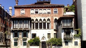 A historic building with a central arched window, a balcony with a wrought iron railing, and a small boat docked in front. - Lovely and quite apartment with garden in the heart of Venice (Venice)