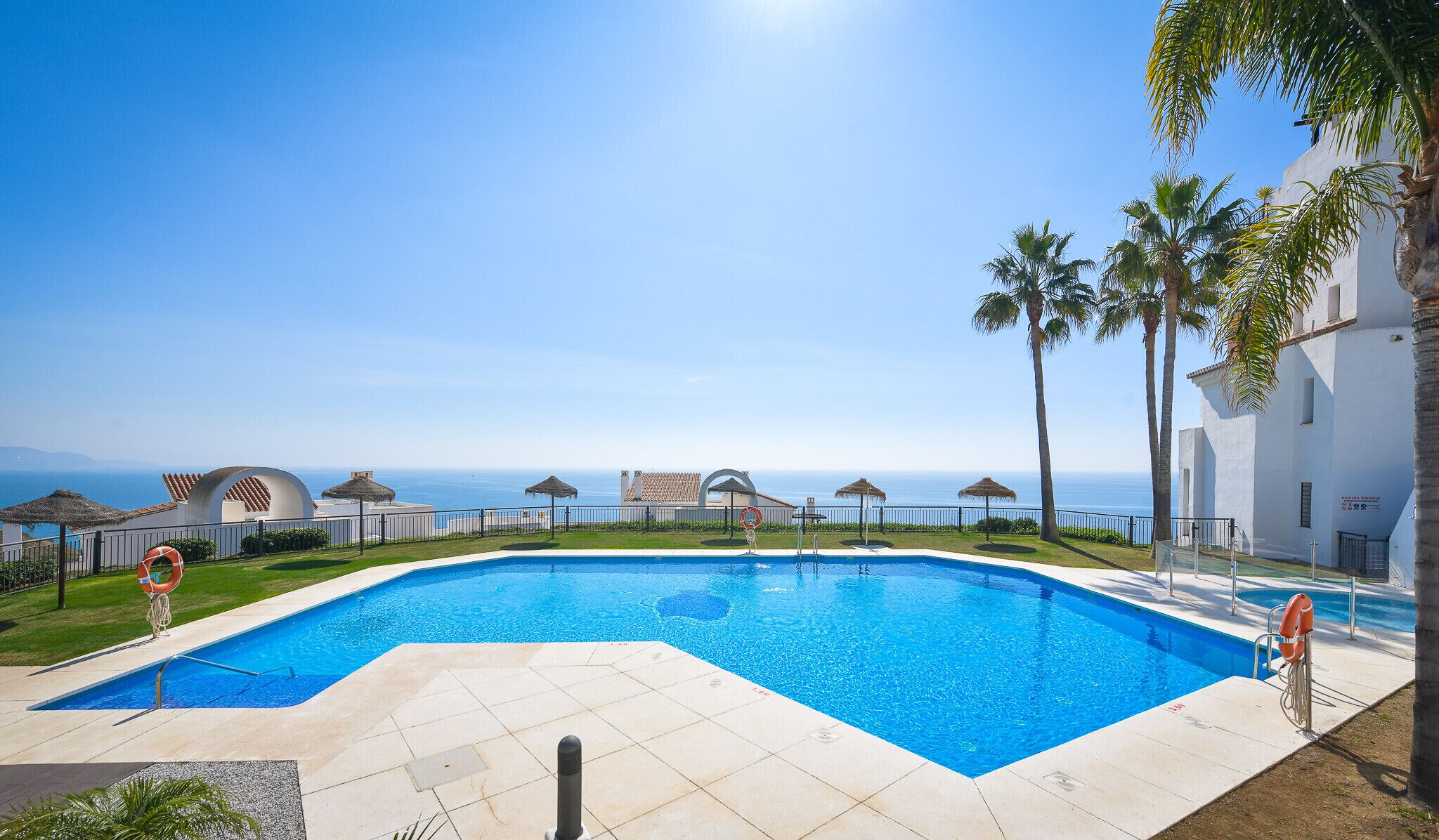 A swimming pool with a clear view of the ocean, surrounded by palm trees and a white building with a sign.