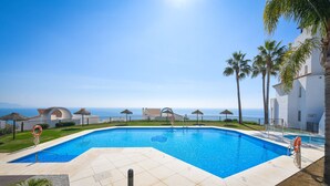 A swimming pool with a clear view of the ocean, surrounded by palm trees and a white building with a sign.