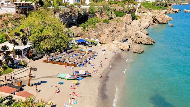 A beach with rocky cliffs, a clear blue sea, and buildings in the background.