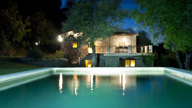 Una casa de pedra amb una piscina, envoltada d'arbres i il·luminada per llums exteriors.