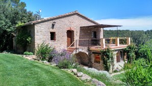 Una casa de pedra amb un balcó, envoltada de vegetació i un cel serè.