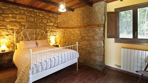 Un dormitori amb paret de pedra, sostre de fusta, llit amb estructura metàl·lica, una petita taula de nit, una làmpada i una finestra amb vista a arbres.
