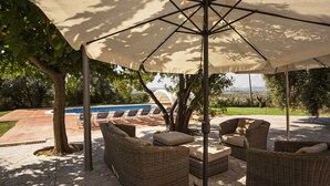An outdoor seating area with wicker furniture and a large umbrella, overlooking a pool and landscaped garden.