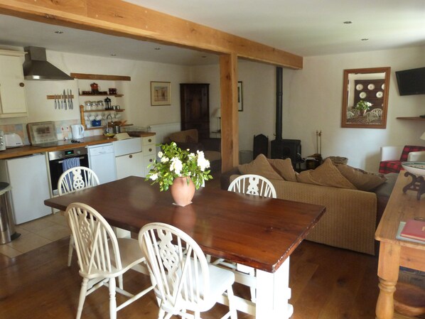 A spacious living area with a wooden dining table, white chairs, and a beige sofa. A kitchen with modern appliances and a large window is visible in the background.