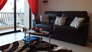 A living room with a black leather sofa, patterned cushions, a glass coffee table with a lace doily, and a red curtain.