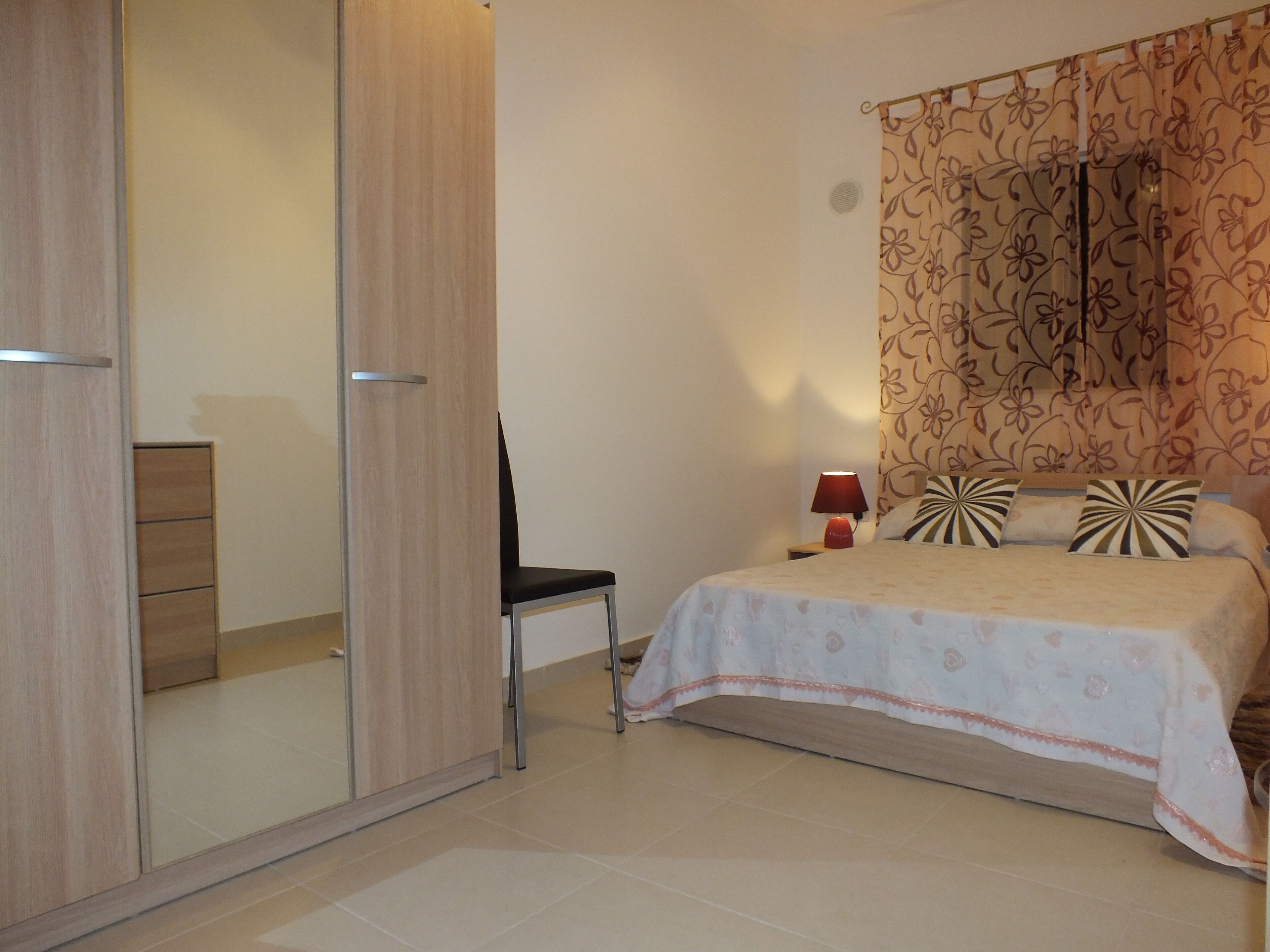 A bedroom with a bed, a chair, a wardrobe, and a nightstand with a lamp.