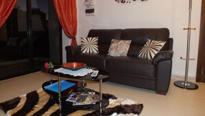 A living room with a dark brown sofa, two patterned throw pillows, a glass coffee table with magazines, a small decorative bowl, and a red curtain.