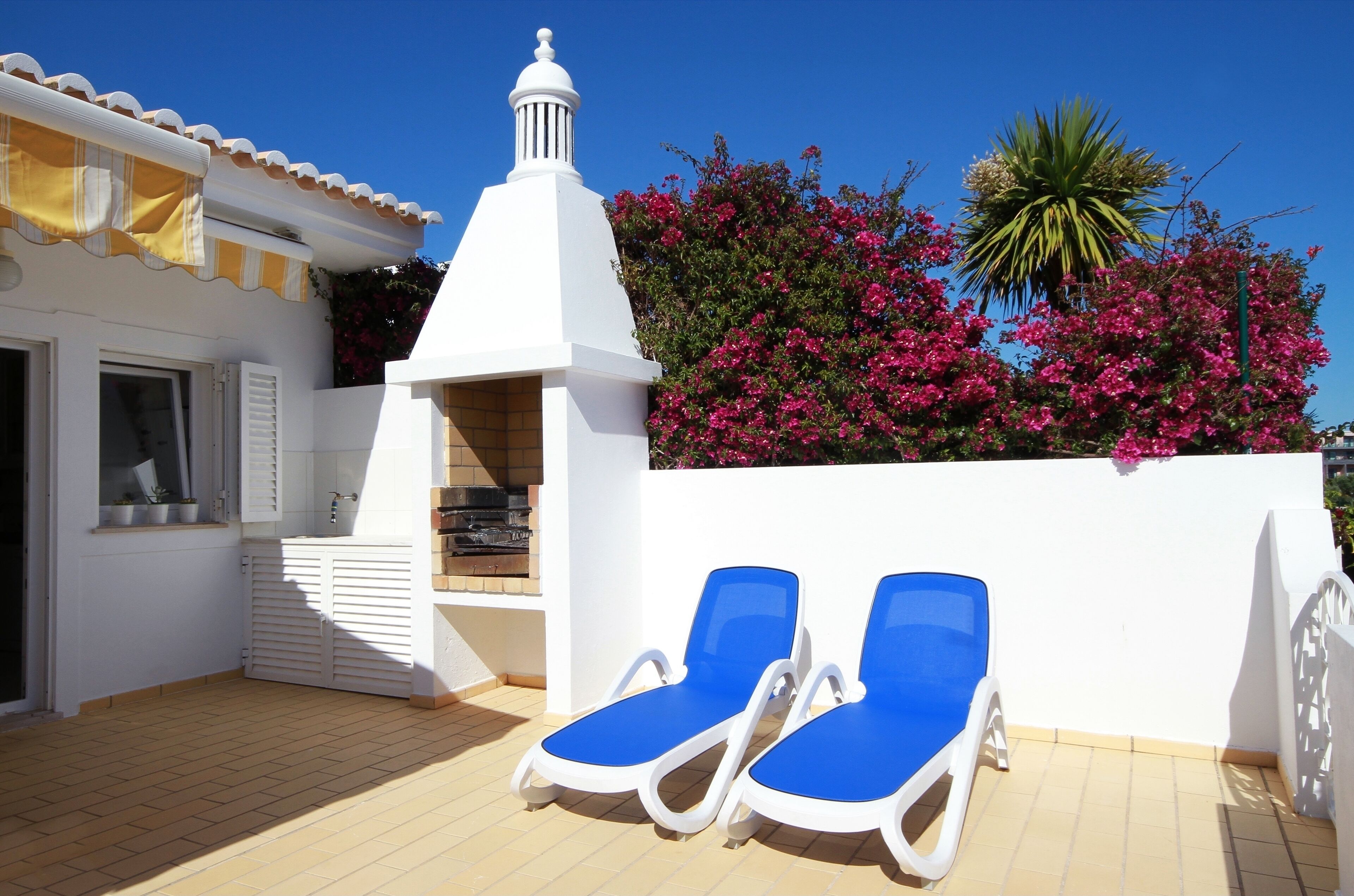 Una terraza en la azotea con dos sillones azules, una chimenea blanca y flores de buganvilla rosadas.