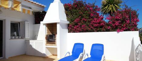 Una terraza en la azotea con dos sillones azules, una chimenea blanca y flores de buganvilla rosadas.