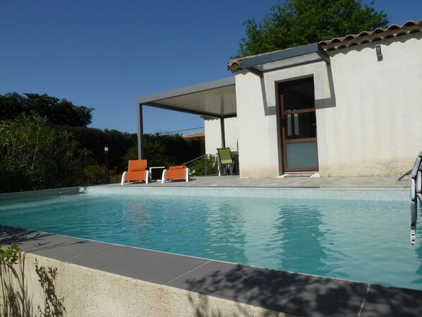 Une piscine, deux chaises longues orange, un patio couvert et un bâtiment doté d’un toit en tuiles.
