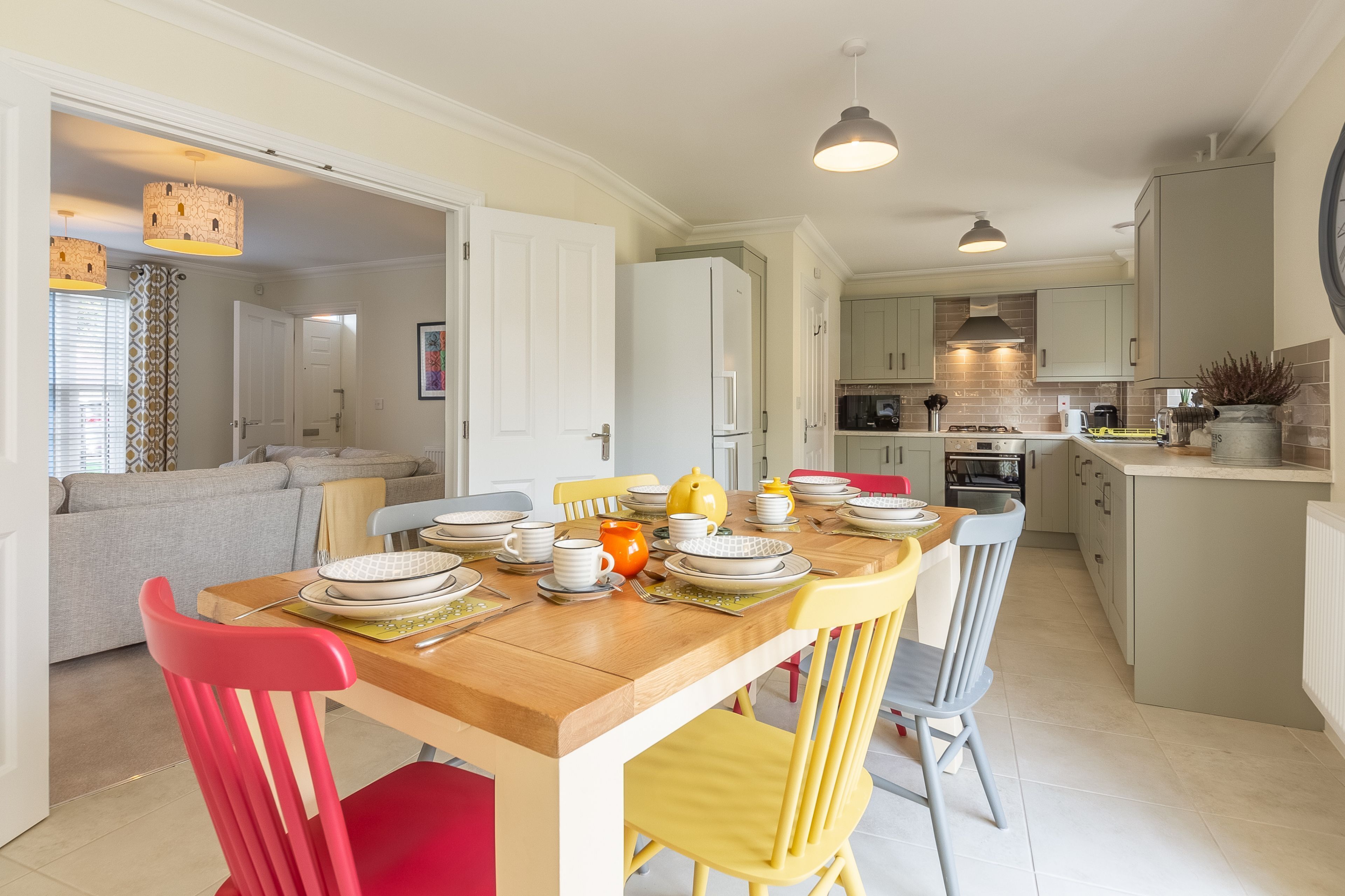 A modern kitchen with a central island, a dining area with a wooden table and chairs, and a living room with a sofa and a patterned curtain.