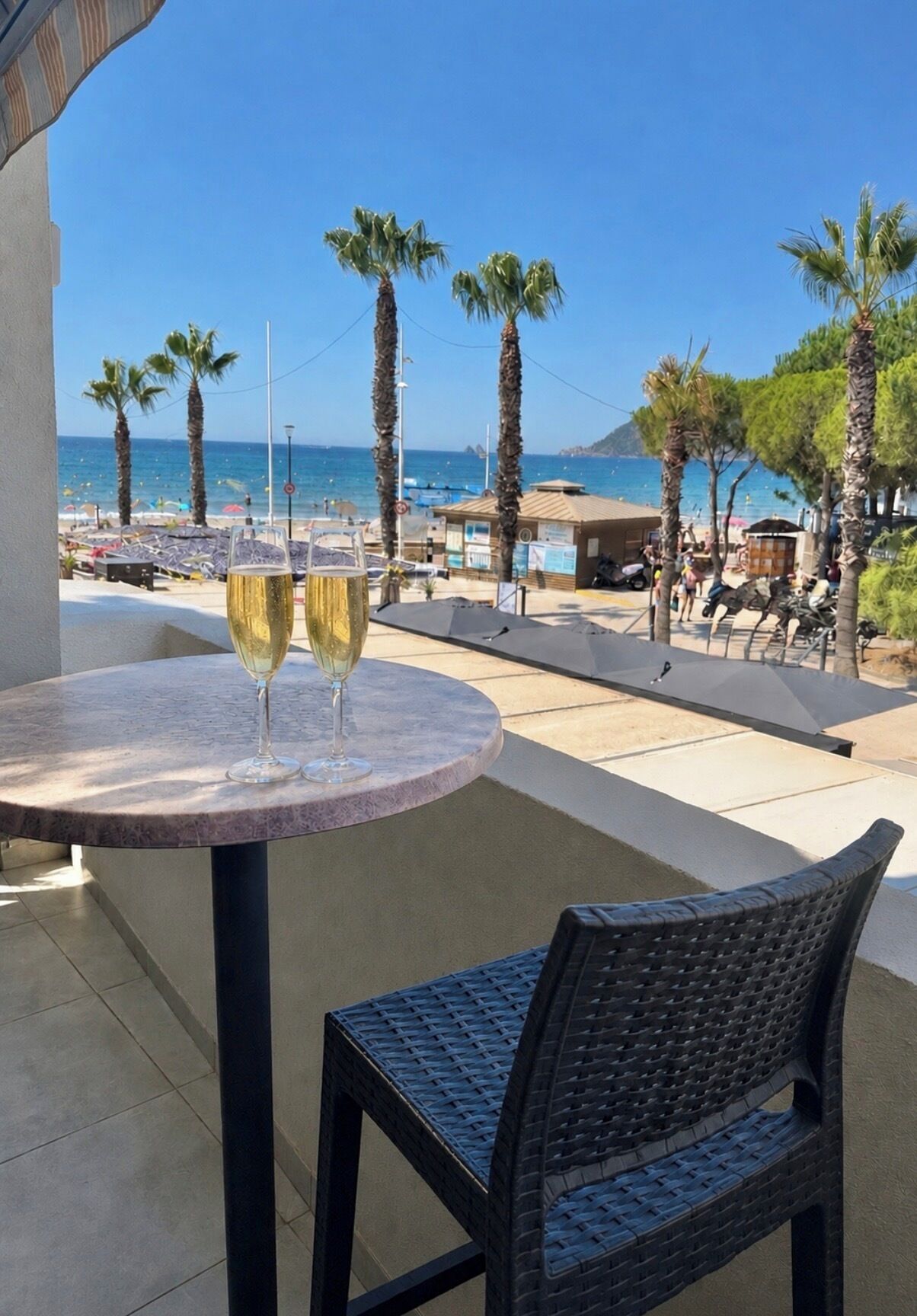 A balcony with a table and two glasses of wine, overlooking a beach with palm trees and the sea.