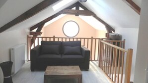 Ein Loft-Zimmer mit einer dunkelgrauen Couch und einem Holztisch. Ein Holzaplicht und ein Fenster mit Runddesign sind zu sehen.